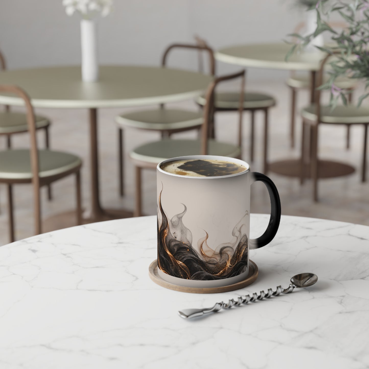 A ceramic coffee mug with a black handle and a marbled smoke design in black, brown, and gold on a cream background, sitting on a marble table in a cafe setting.